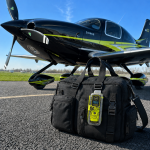 Personal locator beacon ACR ResQLink View RLS fixé au sac de vol d'un pilote à côté d'un avion Cirrus SR22.